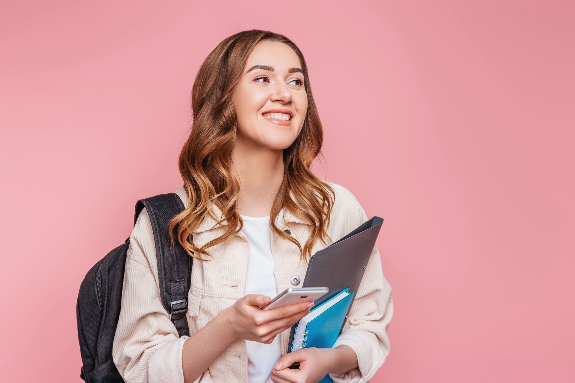 Girl student smiling and looking away holding a smartphone in her hands isolated on a pink background