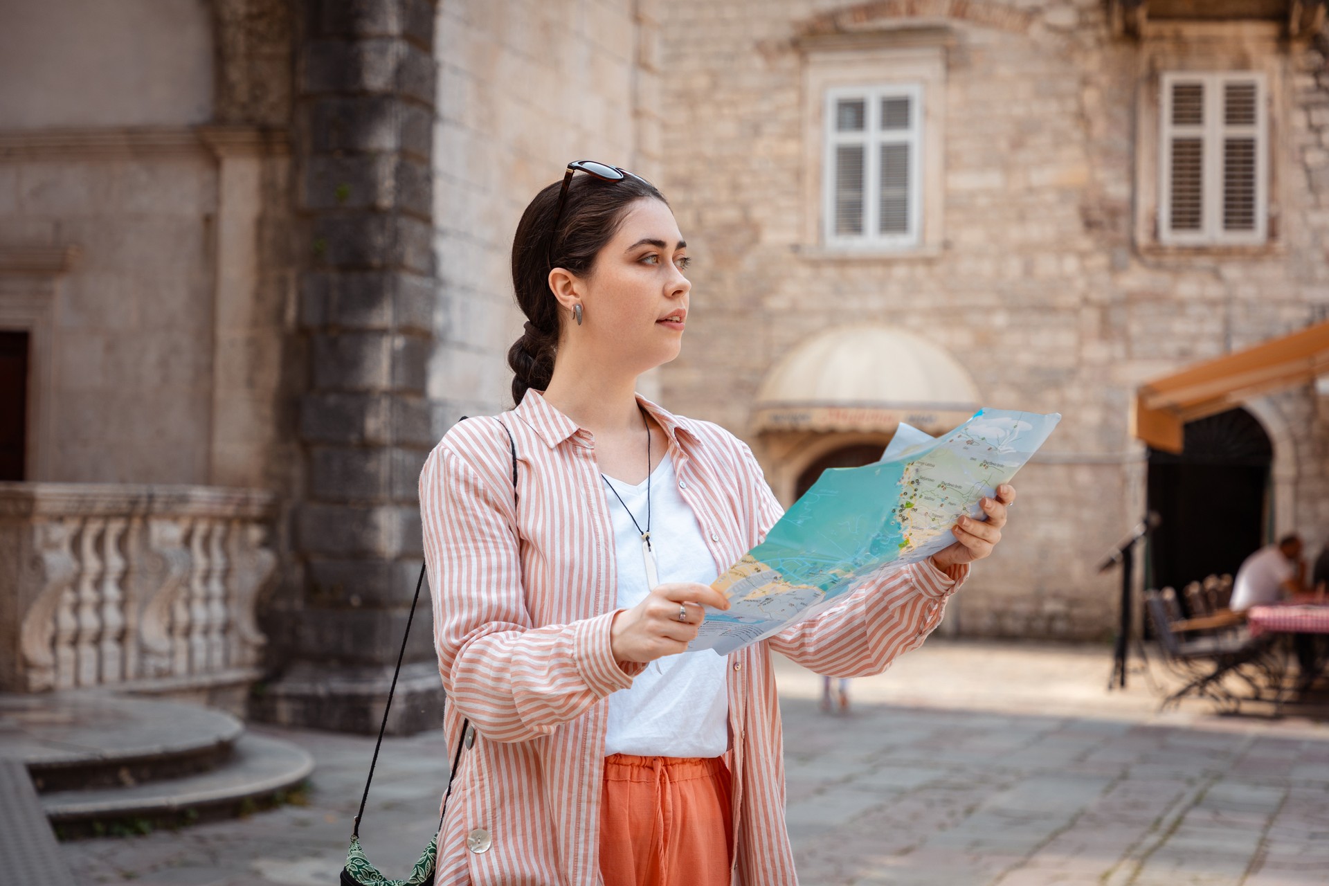 Young pretty Caucasian woman holds a paper map in her hand and walks around an old European city for sightseeing. Summer vacation alone